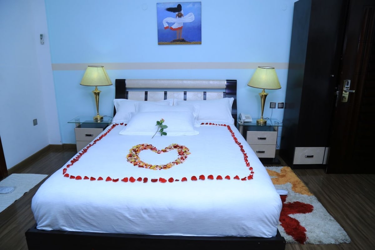 A neatly made bed with a heart-shaped flower arrangement, two bedside lamps, a painting on the wall, and a door to the right.