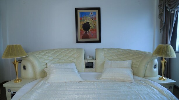A double bed with a quilted headboard, bedside lamps, a small bedside table, a framed artwork on the wall, and a window with curtains.
