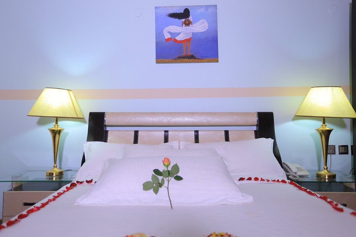 A hotel room with a bed, two bedside lamps, a painting on the wall, and a single rose on the pillow.