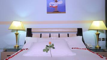 A hotel room with a bed, two bedside lamps, a painting on the wall, and a single rose on the pillow.