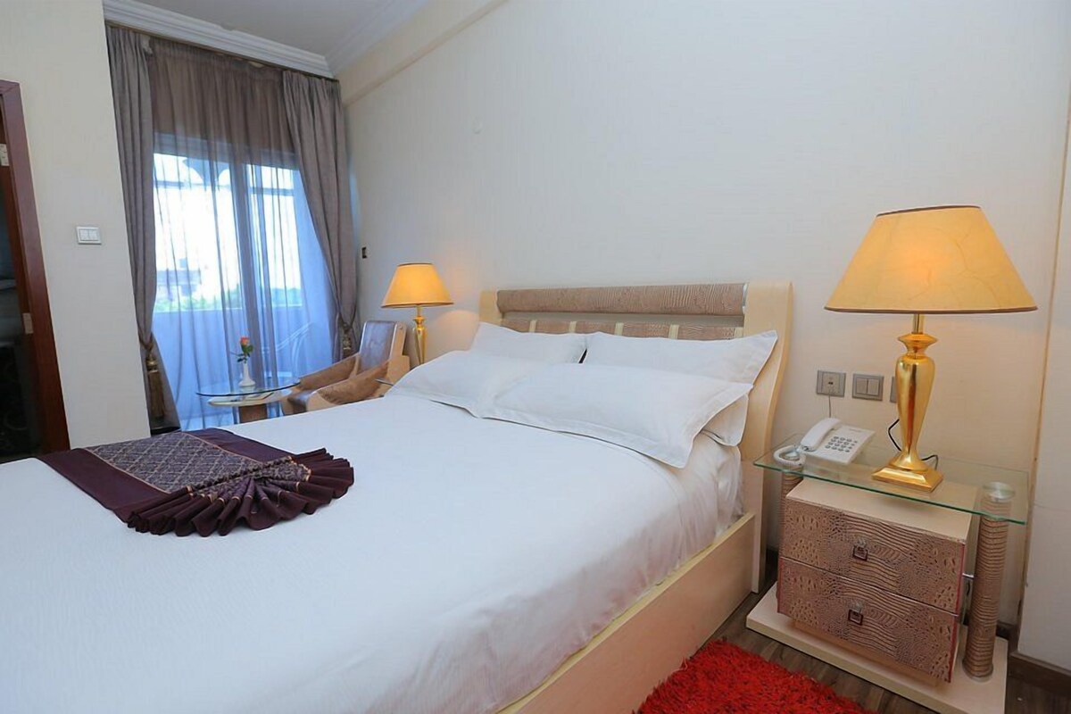 Una habitación de hotel con una cama, lámparas de noche, una mesita de vidrio con un teléfono y una alfombra roja.
