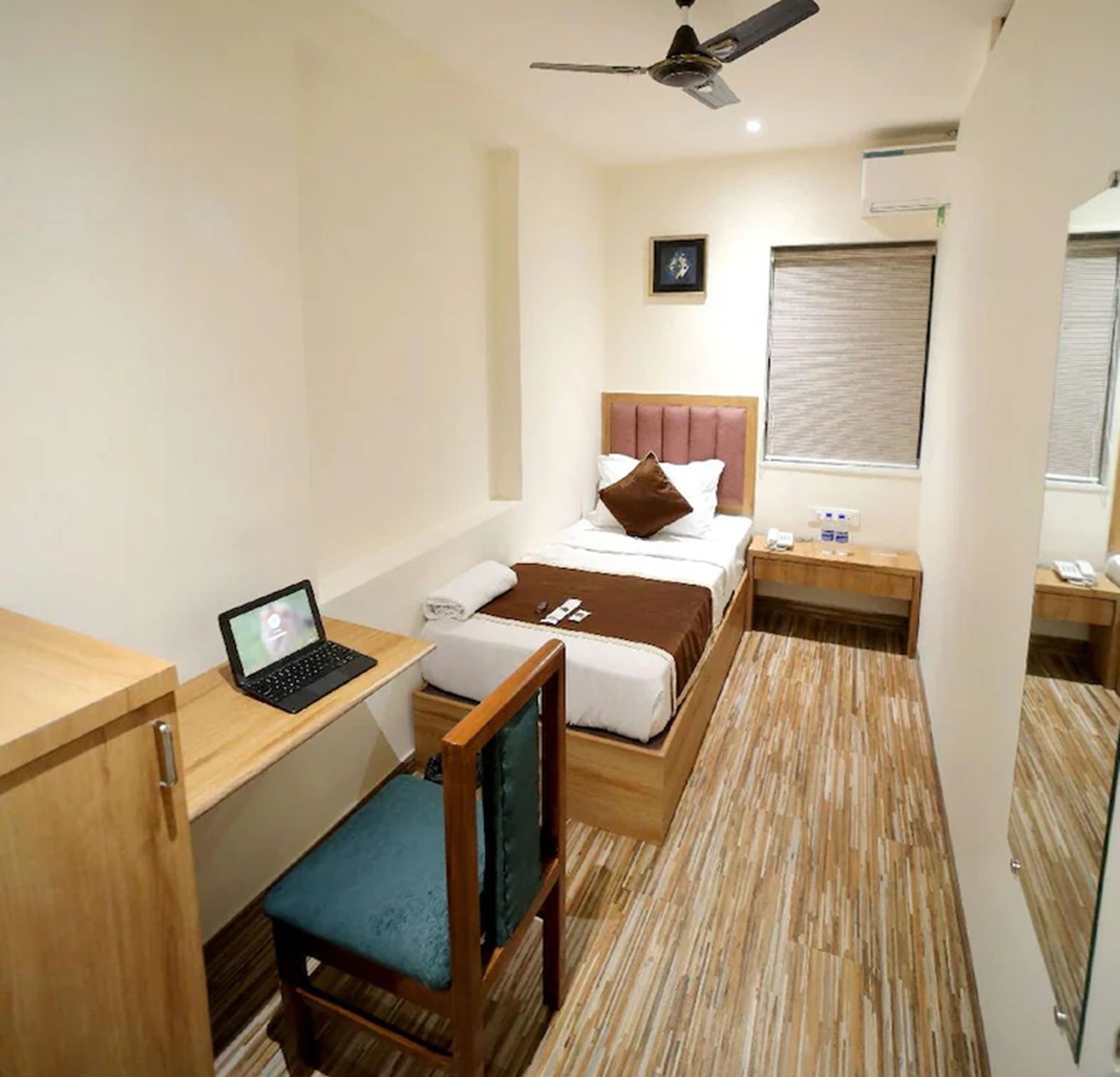 A compact hotel room with a bed, a desk with a laptop, a chair, a bedside table, and a ceiling fan.