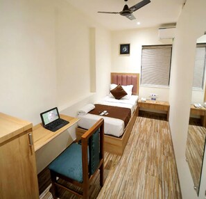 Ein kleines Hotelzimmer mit einem Bett, einem Schreibtisch mit Laptop, einem Sessel, einem Nachttisch und einem Deckenventilator.