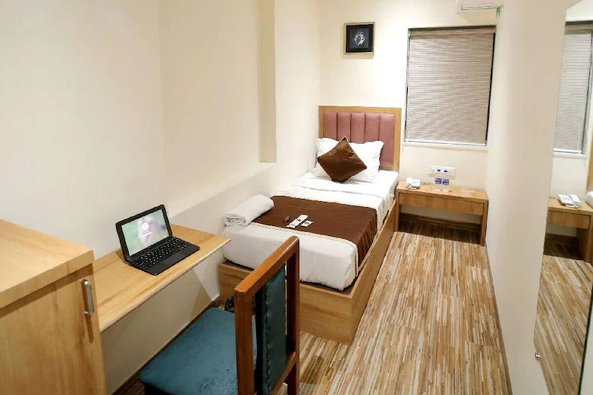 A compact hotel room with a bed, a desk with a laptop, a chair, a bedside table, and a ceiling fan.