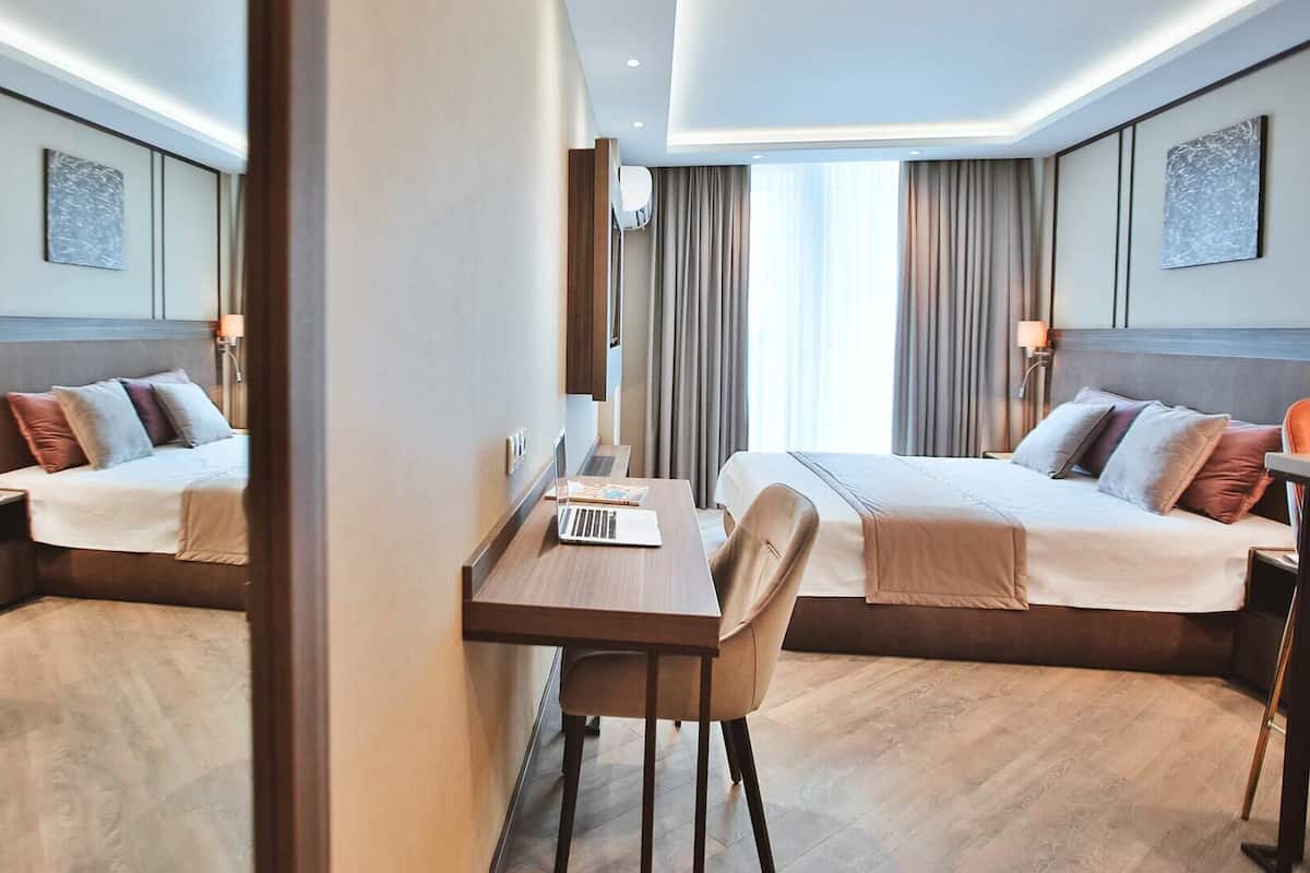 Hotellihuoneessa on kaksi sänkyä, työpöytä ja tuoli. Työpöydällä on kannettava tietokone, ja huoneen sisustus on moderni ja värit ovat hillittyjä.