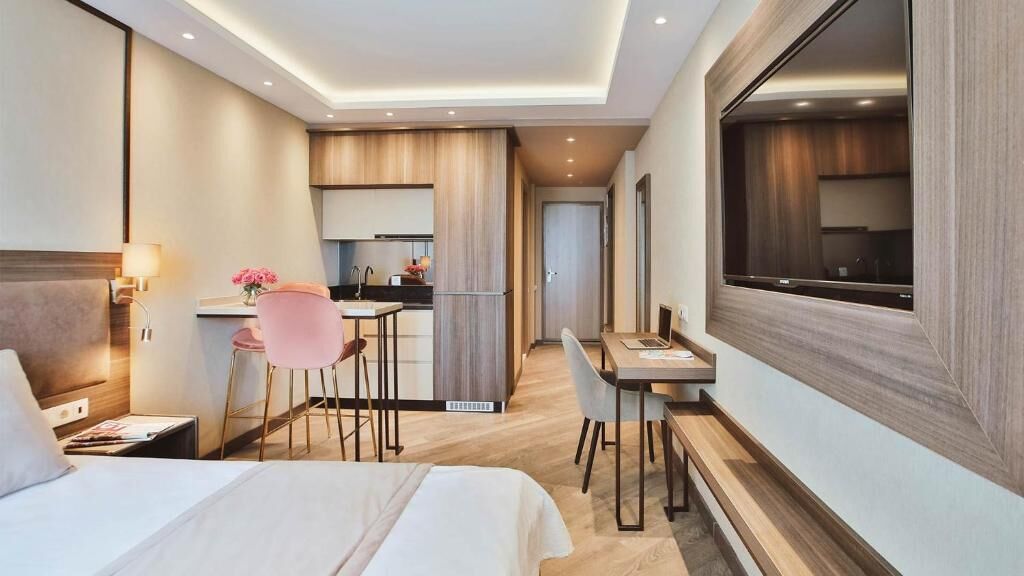 Moderni hotellihuone, jossa on suuri sänky, työpöytä, tuoli ja pieni keittiönurkkaus.