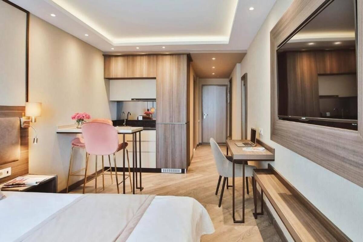 A modern hotel room with a large bed, a desk, a chair, and a kitchenette.