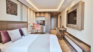Ein Hotelzimmer mit Bett, Schreibtisch, Stuhl, Fernseher und einer kleinen Küche im Hintergrund.