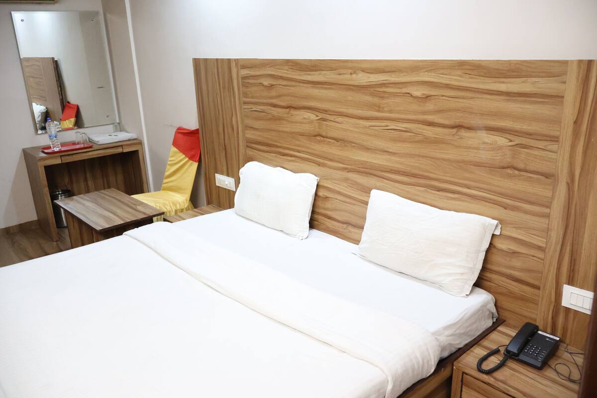 A hotel room with a wooden headboard, a bed with white bedding, a bedside phone, a small wooden table with a chair, and a mirror above the sink.