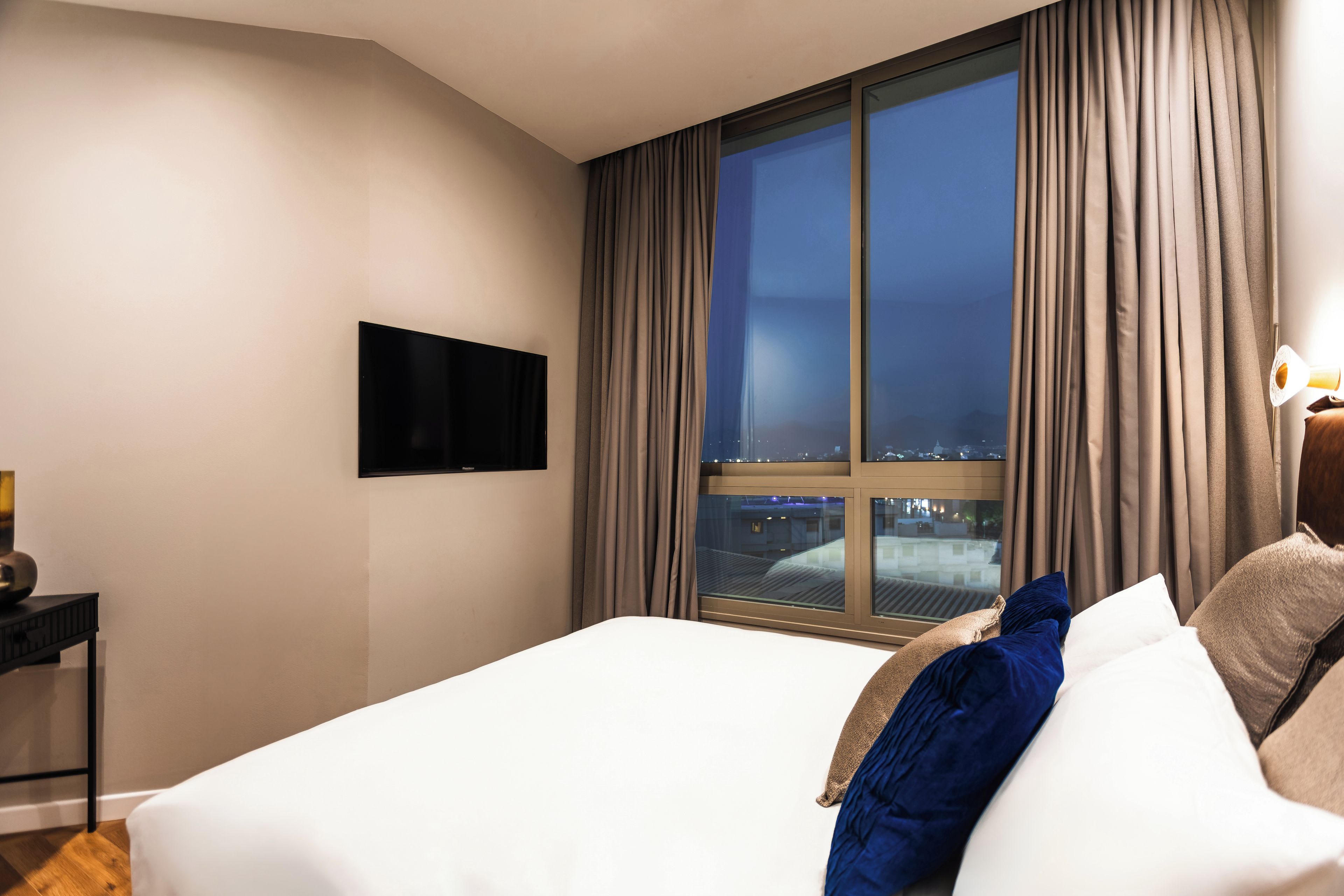 A hotel room with a large bed, a flat-screen TV mounted on the wall, and a window with curtains.