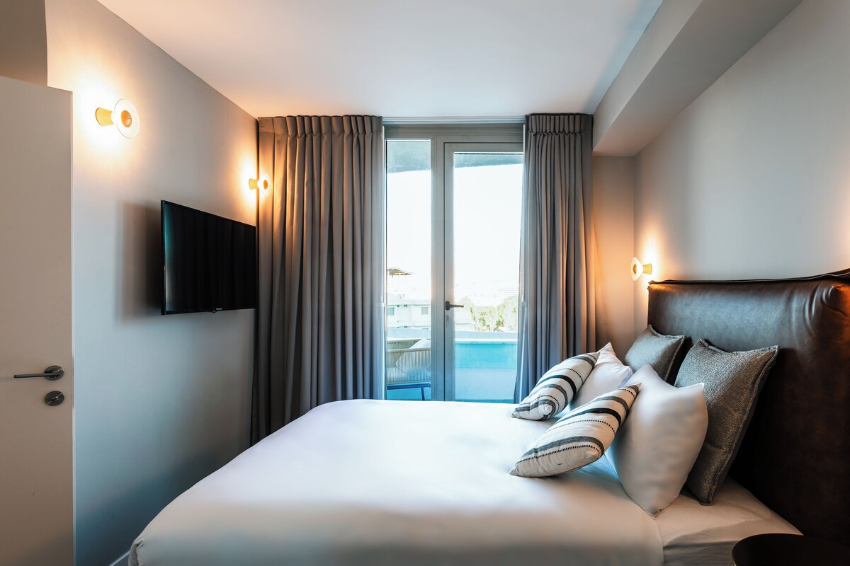Habitación de hotel con cama, televisor montado en la pared y vista a una piscina por la ventana.