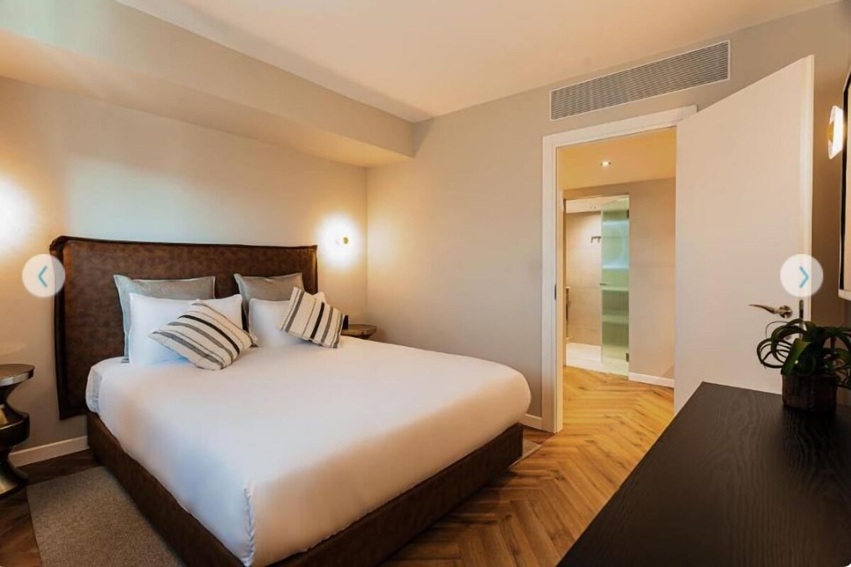 A bedroom with a bed, bedside tables, a nightstand, a wooden floor, and a door leading to another room.