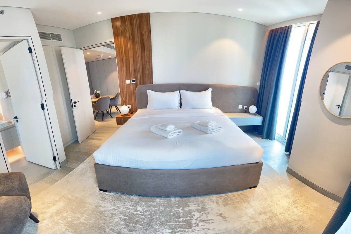 A modern hotel room with a large bed, a seating area, a dining table, and a bathroom visible through an open door.