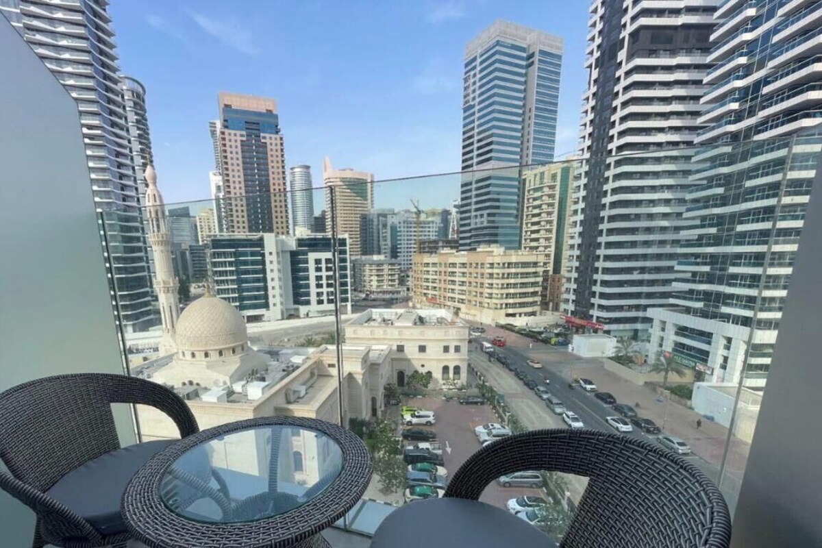 A balcony with wicker chairs and a glass table overlooking a cityscape with modern buildings and a dome structure.