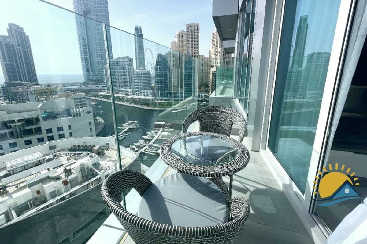 A balcony with wicker chairs and a glass table overlooking a cityscape with high-rise buildings and a marina.