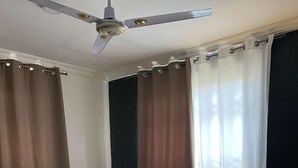 Quarto com cama, cortinas, ventilador de teto, mesa de cabeceira com luminária e piso estampado.