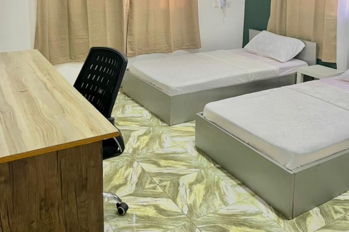 A room with two beds, a desk, a chair, and a ceiling fan.