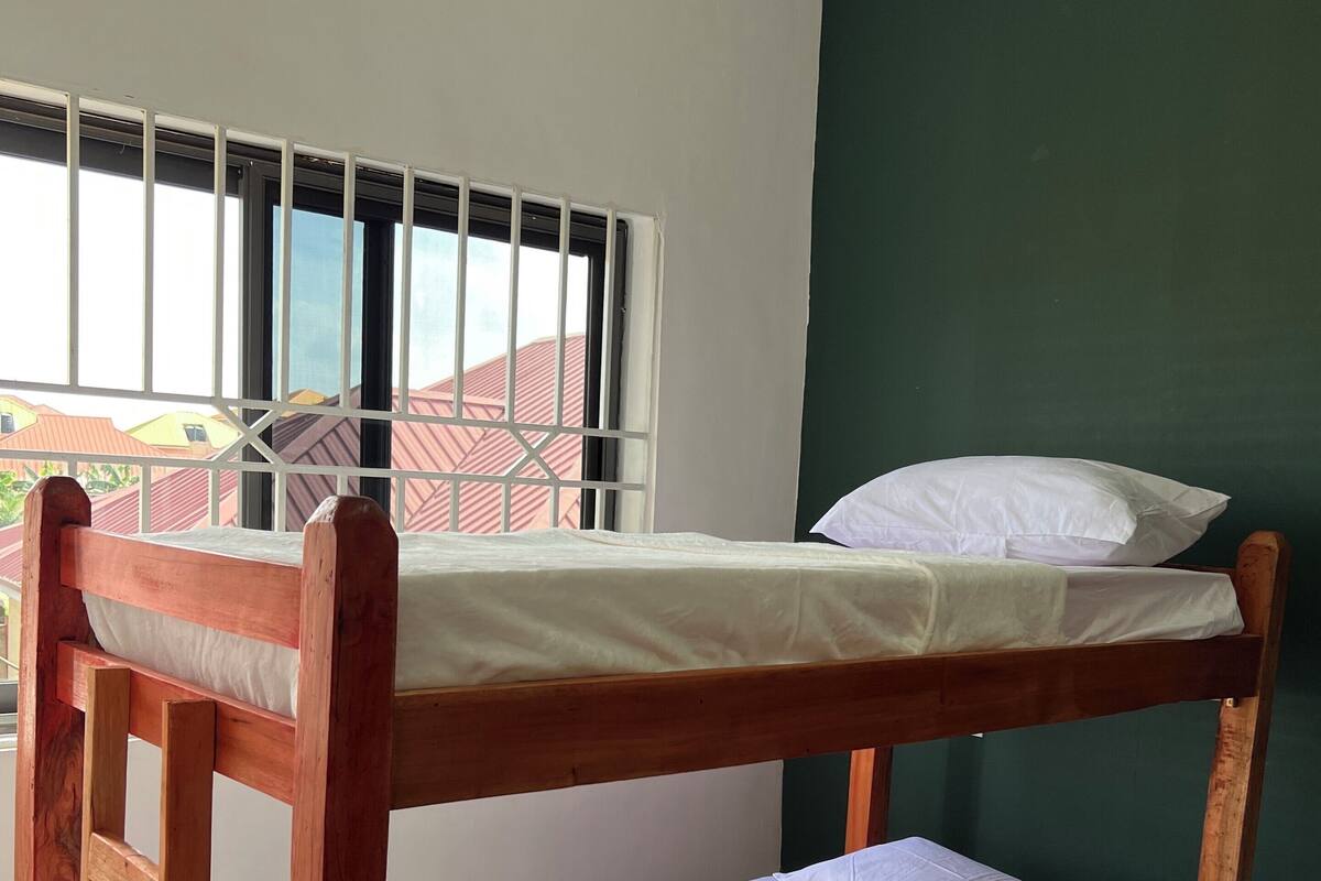 Una cama litera con ropa de cama blanca en una habitación con pared verde y una ventana con barrotes.