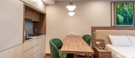 A compact room with a kitchenette, a dining table with chairs, a bed, and a painting on the wall.