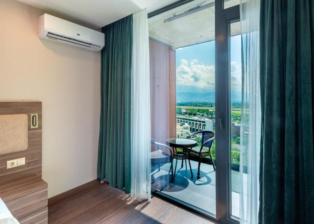 Una habitación con cama, aire acondicionado, balcón con mesa y sillas, y vista a la ciudad.