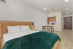 Ein modernes Hotelzimmer mit einem Holzkopfteil, einer grünen Bettdecke, einer kleinen Küchenzeile und einer Tür zu einem weiteren Zimmer.