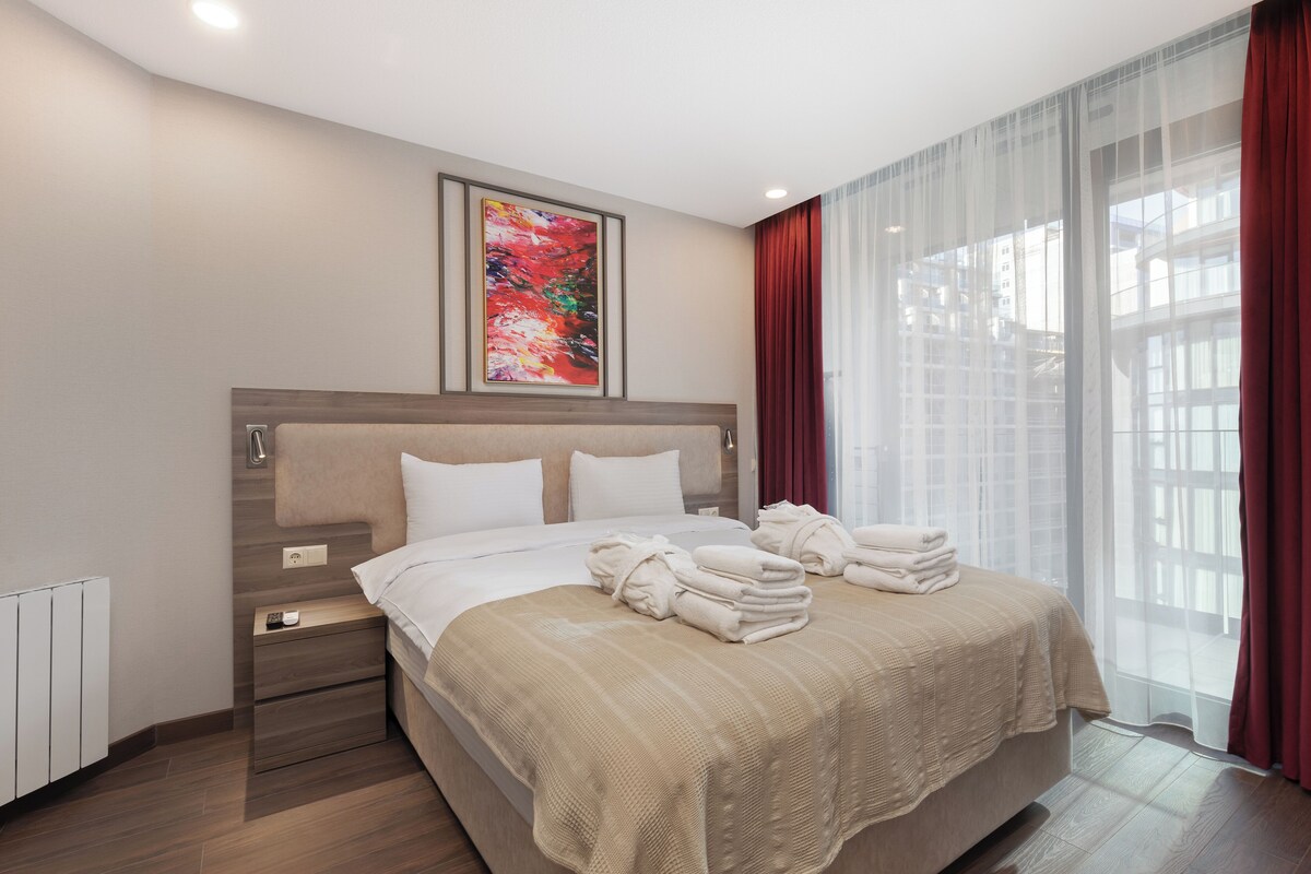 Ein modernes Hotelzimmer mit einem großen Bett, einem Nachttisch, einem Fenster mit Vorhängen und einem Bild an der Wand.