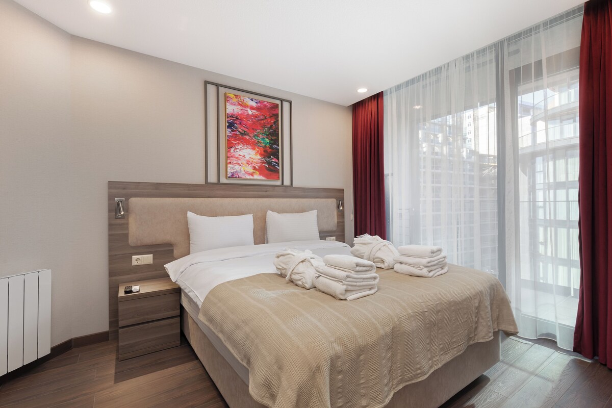 Ein modernes Hotelzimmer mit einem großen Bett, einem Nachttisch, einem Fenster mit Vorhängen und einem Bild an der Wand.