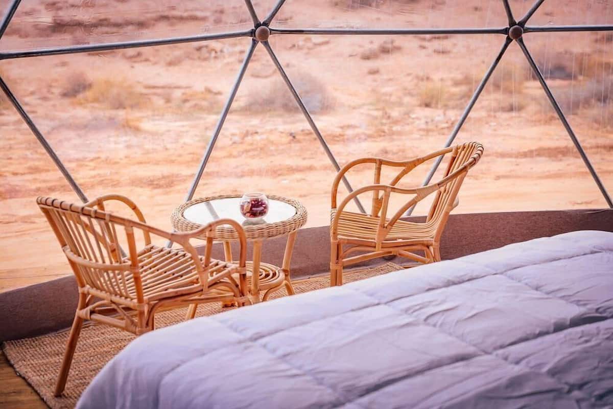 Um quarto acolhedor com uma cama, duas cadeiras de vime, uma mesa redonda pequena e vista para uma paisagem desértica através de uma janela geométrica.