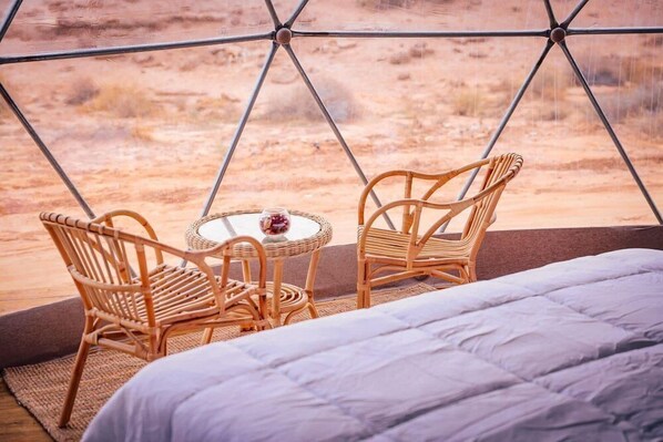 Een knus vertrek met een bed, twee rieten stoelen, een klein rond tafeltje en uitzicht op een woestijnlandschap door een geometrisch raam.