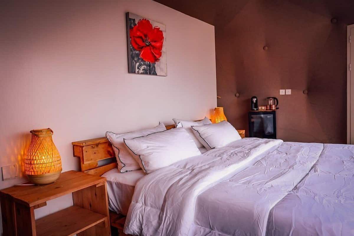 Quarto com cama, almofadas, mesa de cabeceira, luminária, cafeteira e um quadro na parede.