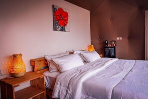 A bedroom with a bed, pillows, a nightstand, a lamp, a coffee maker, and a painting on the wall.