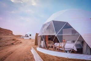 Een woestijnlandschap met een grote geodetische koepelstructuur, daarbinnen een bed en stoelen.