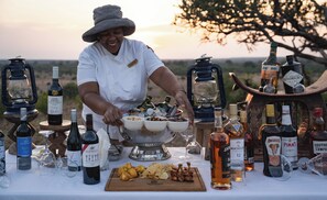 A person serving food at an outdoor event with a variety of bottles and glasses on a table. - Mashatu Lodge (Lentswelemoriti)