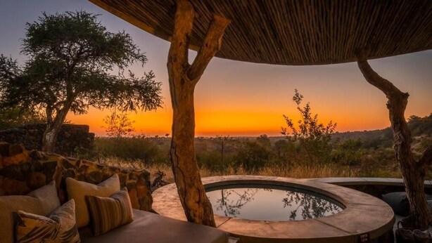Un espacio circular de estar al aire libre con una piscina, rodeado de muros de piedra y árboles, con vista a un atardecer.