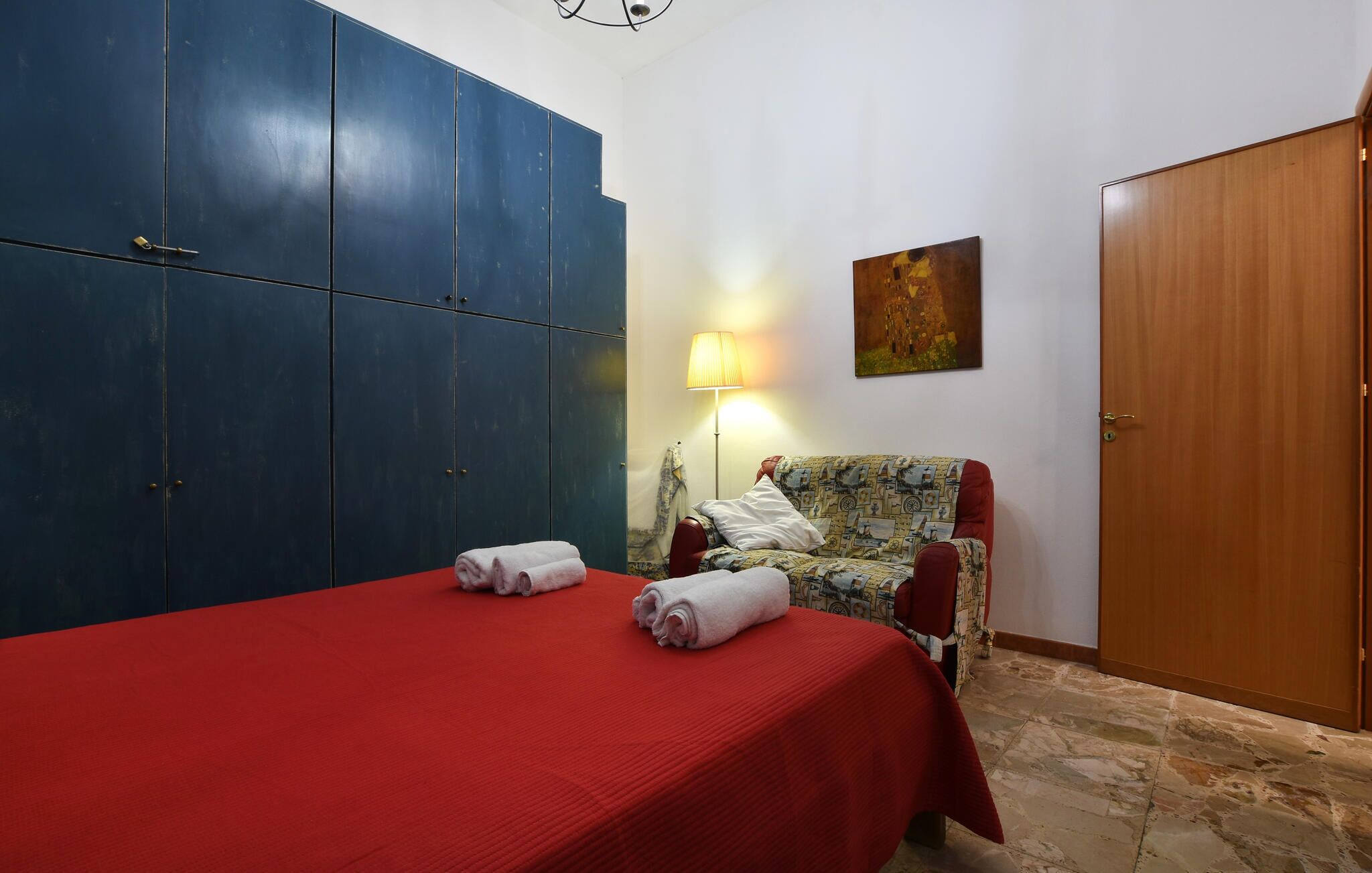 A room with a bed, a chair, a wardrobe, a lamp, and a painting on the wall.
