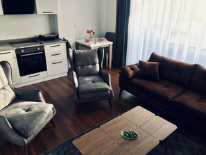 Ein modernes Wohnzimmer mit einem braunen Sofa, grauen Sesseln, einem Holztisch und einer Küchenzeile mit weißen Schränken und einem Einbauofen.