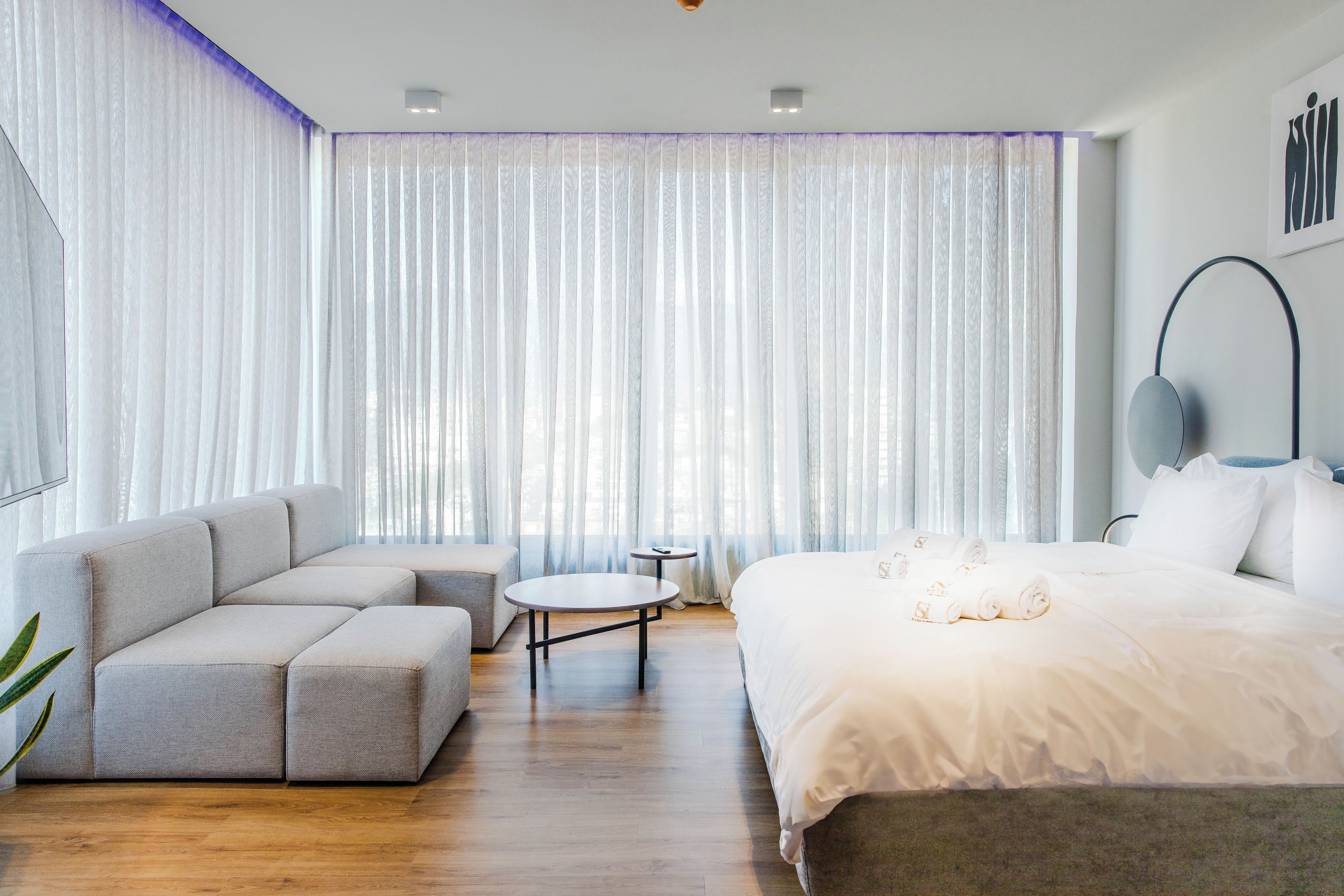 A modern hotel room with a sectional sofa, a round coffee table, a large bed, and a small side table.