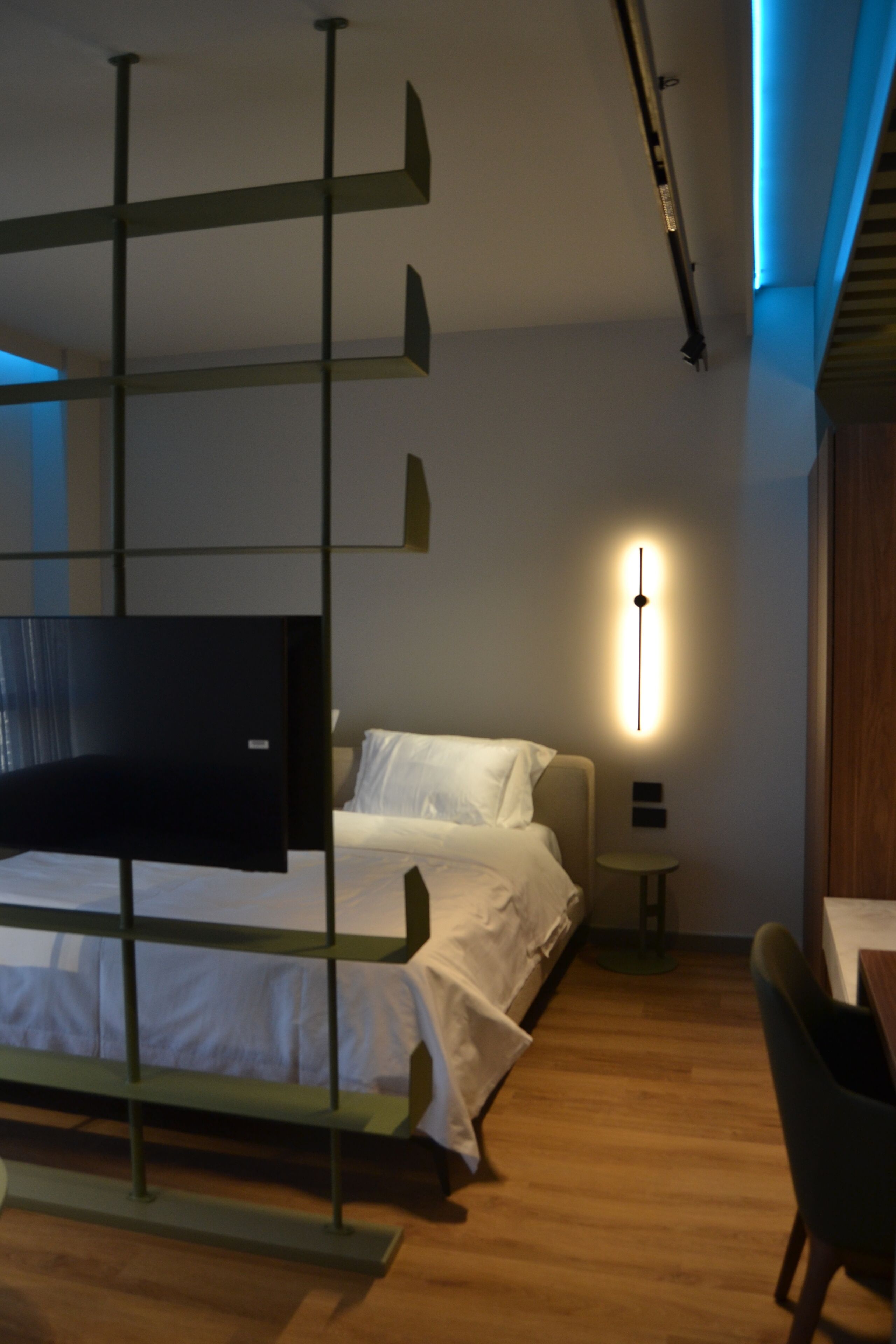 A modern hotel room with a bed, a desk, a chair, a TV, and a wall-mounted light fixture.