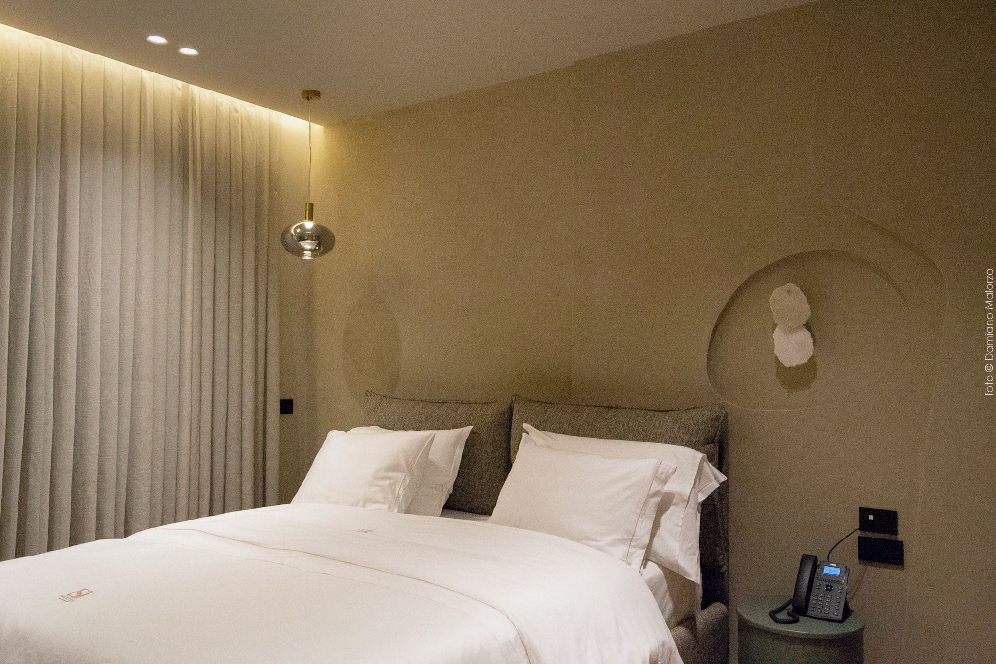 Una habitación de hotel con una cama, una mesita de noche con teléfono, una lámpara de pared y una cortina.