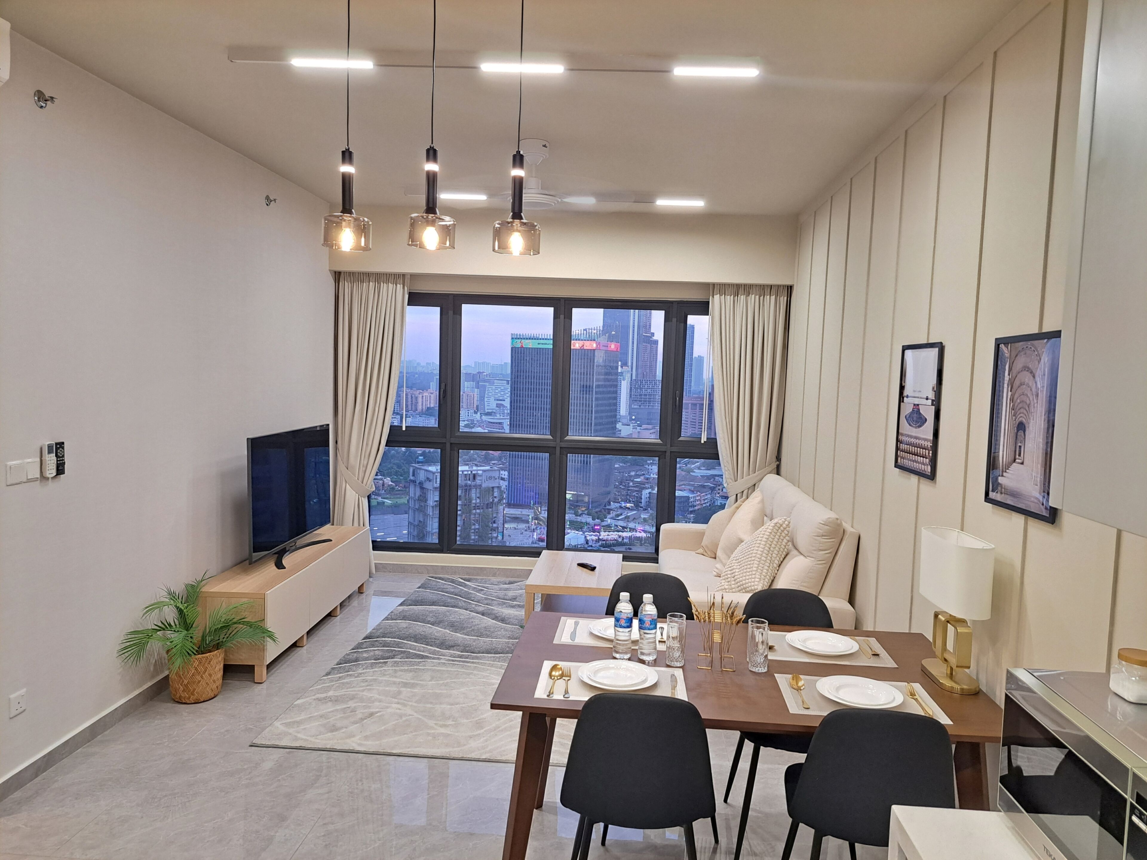 A modern living room with a dining area, large windows with a city view, a flat-screen TV, and a dining table set for four.