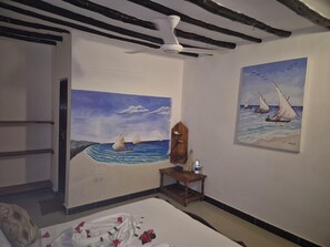 Une chambre à coucher avec un lit, un ventilateur de plafond, un tableau représentant des voiliers et une table de chevet en bois sur laquelle se trouve un vase.