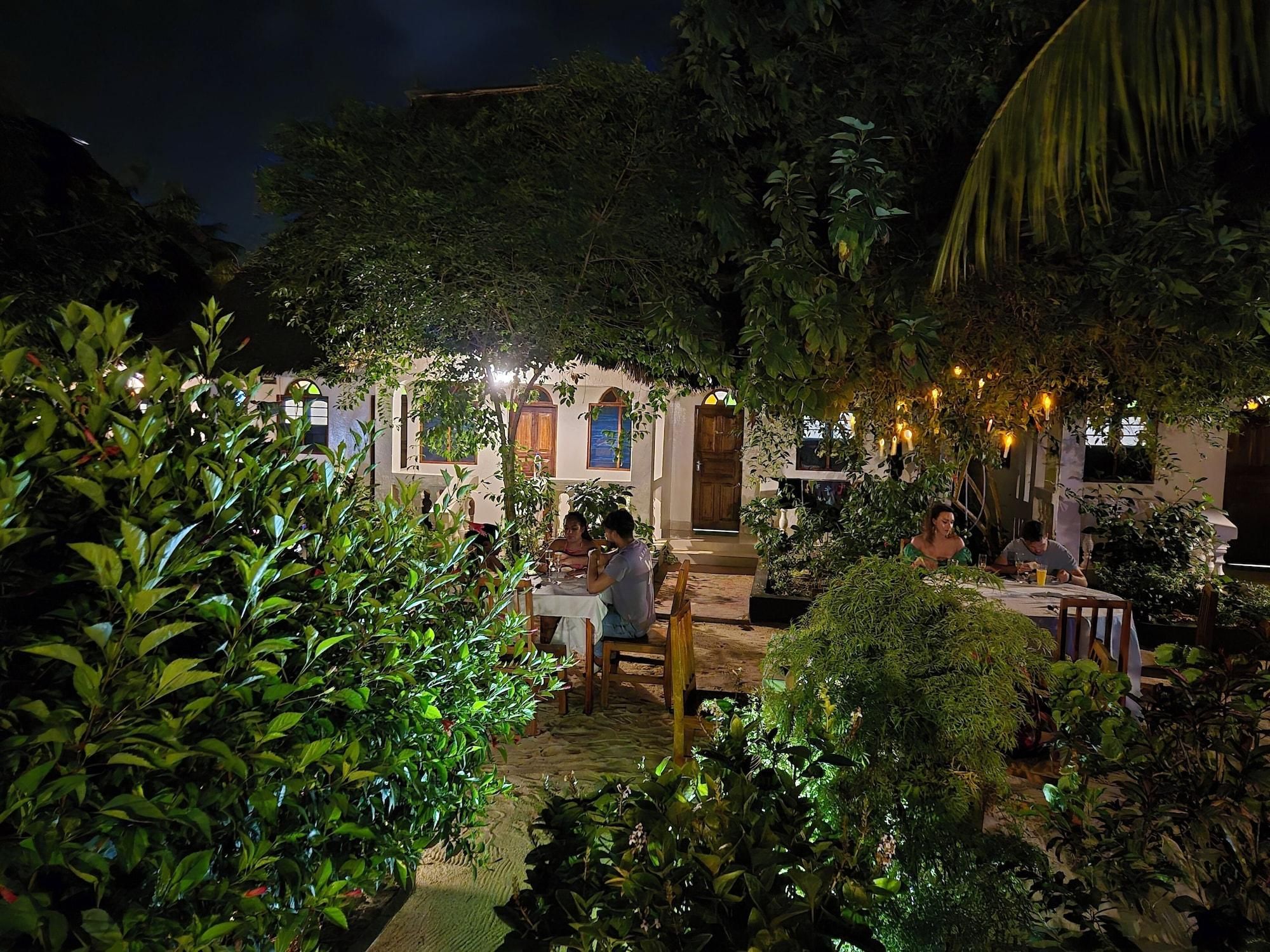 An outdoor dining area with tables and chairs, surrounded by lush greenery and a building with arched windows.