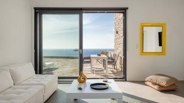 A modern living room with a white sofa, a coffee table with decorative items, a yellow-framed mirror, and a view of the sea through large glass doors. - Ocean Villa Paros (Paros)