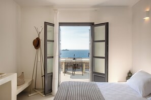 A bedroom with a bed, a nightstand, a window with curtains, and a view of the sea and a table outside. - Ocean Villa Paros (Paros)