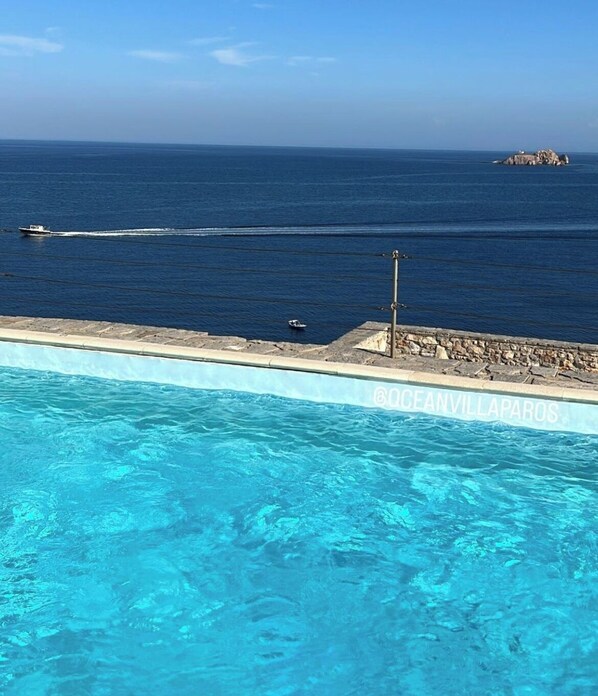 Ein Swimmingpool mit klarem, blauem Wasser, einer Steinmauer und Blick auf das Meer mit Booten.