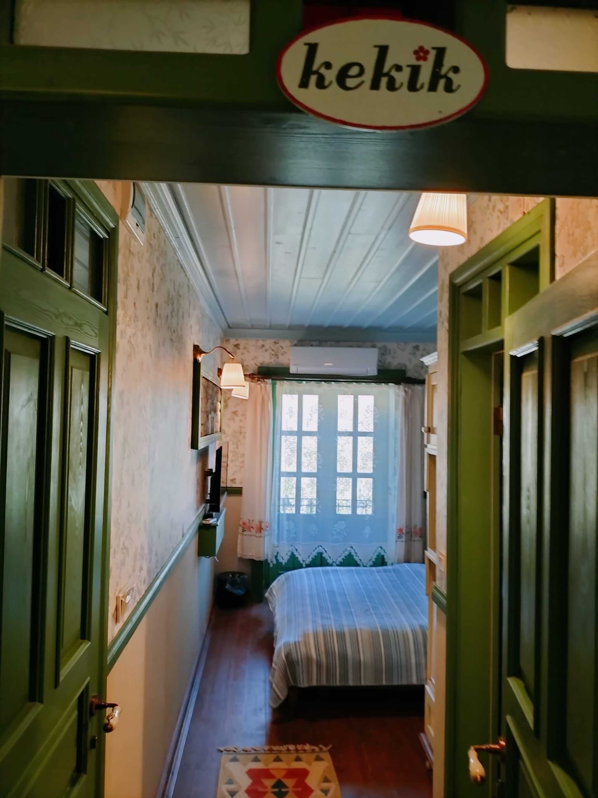 A narrow hallway leading to a room with a bed, a window with lace curtains, and a ceiling fan.