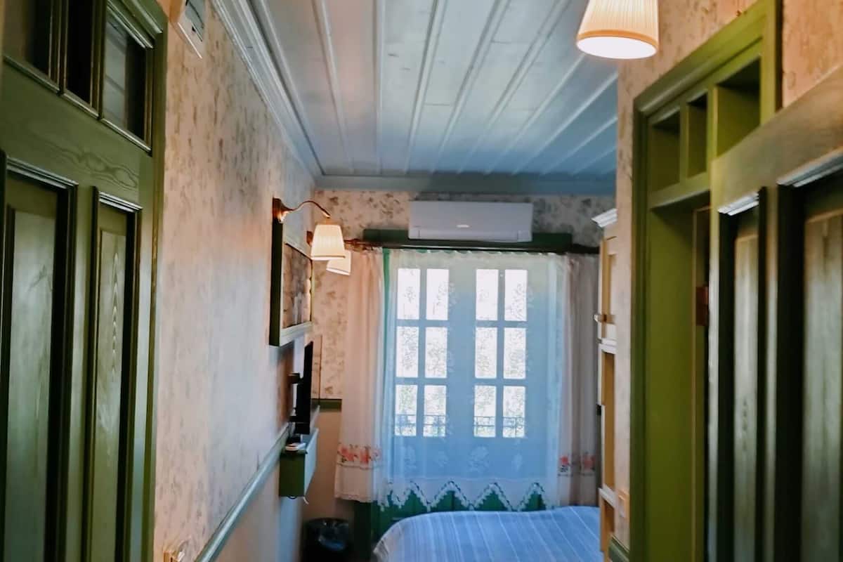 A narrow hallway leading to a room with a bed, a window with lace curtains, and a ceiling fan.