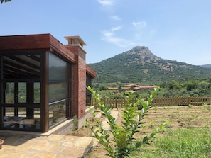 A modern building with large windows and a brick chimney, set against a backdrop of a mountain and a fenced garden area. - Yeşil Ev Butik Otel & Kafe (Gökçeada)