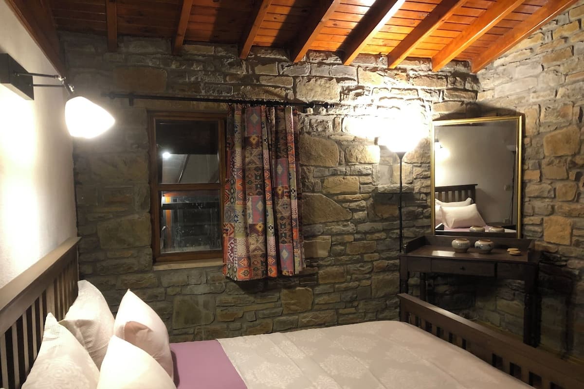 A bedroom with a stone wall, wooden beams, a bed with pillows, a window, and a mirror.