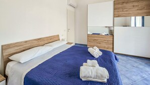 Un dormitori amb un llit, un panell de fusta al capçal, un edredó blau i un coixí blanc. Hi ha un armari de fusta i un mirall a la paret.